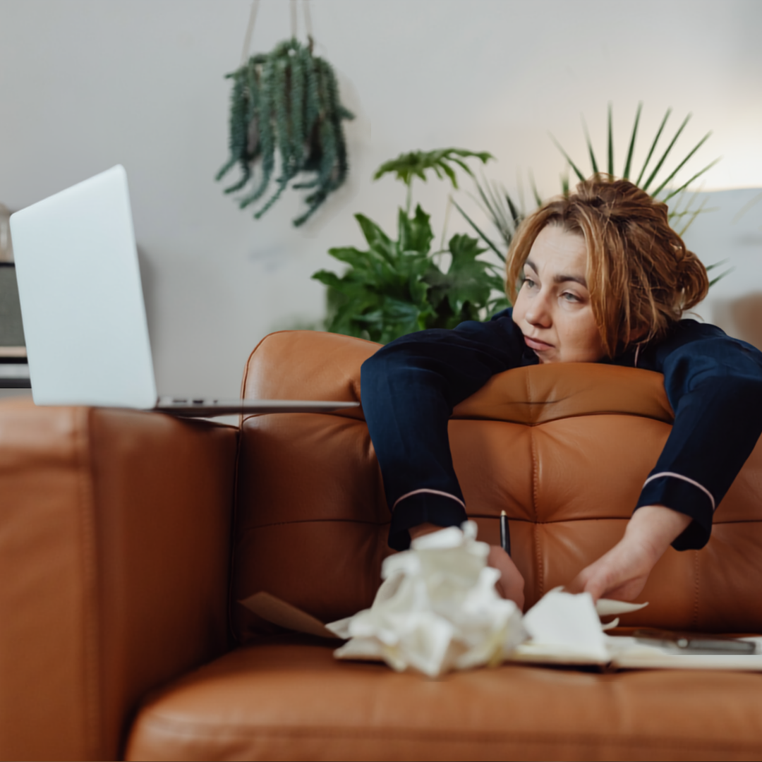 A tired woman in pyjamas slouches on a brown sofa, leaning over the backrest toward a laptop. Used tissues and notebooks are on the table. Green indoor plants are in the background.