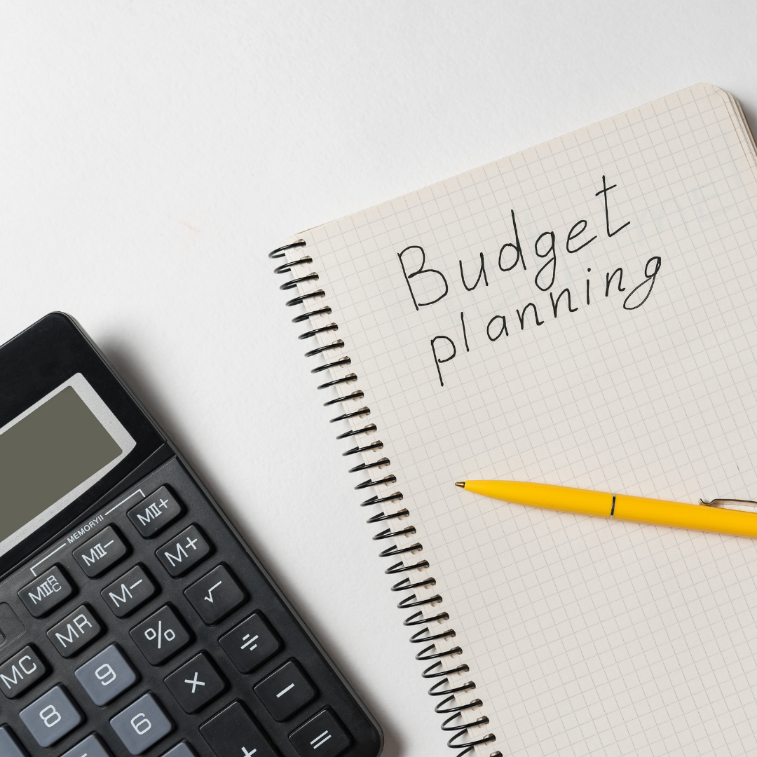 A black calculator, a yellow pen, and a spiral notebook with Budget planning written on the page, all arranged on a white surface.