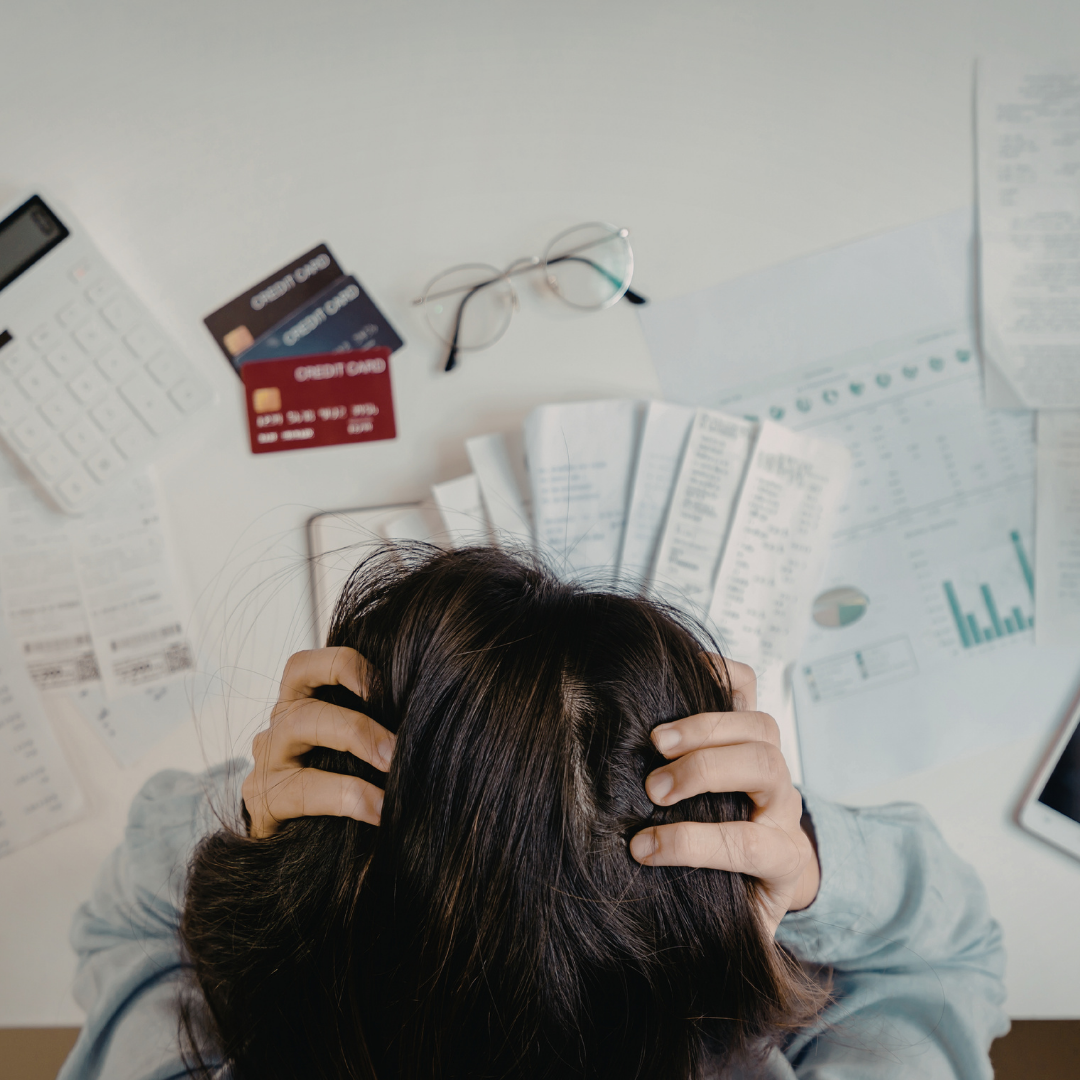 A person holds their head in frustration at a desk covered with receipts, bills, credit cards, a calculator, eyeglasses, and financial documents. The scene suggests financial stress or overwhelm with expenses.