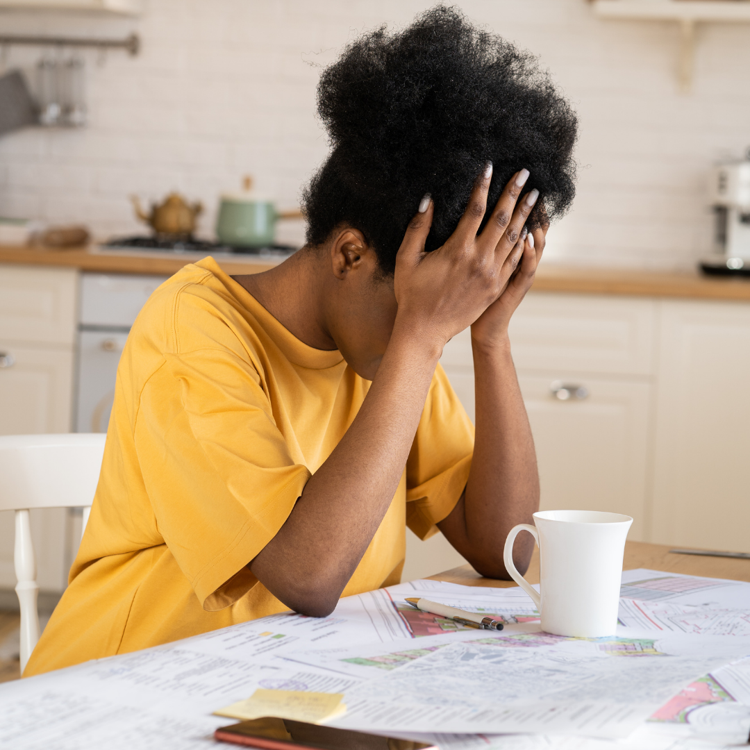 A person sitting at a kitchen table, holding their head in their hands, looking stressed. Papers, documents, and a mug are spread out on the table in front of them.