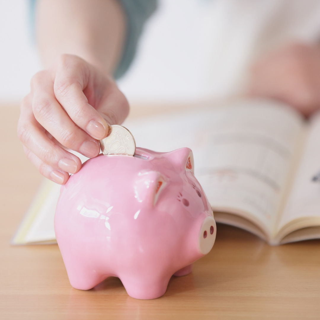 A person places a coin into a pink piggy bank of savings on a wooden table, with an open book in the background.