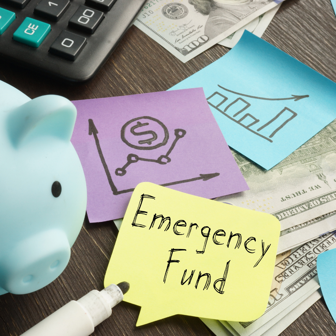 A blue piggy bank, a calculator, dollar bills, and colorful sticky notes showing financial graphs, a dollar sign, and one labeled Emergency Fund are arranged on a wooden surface.