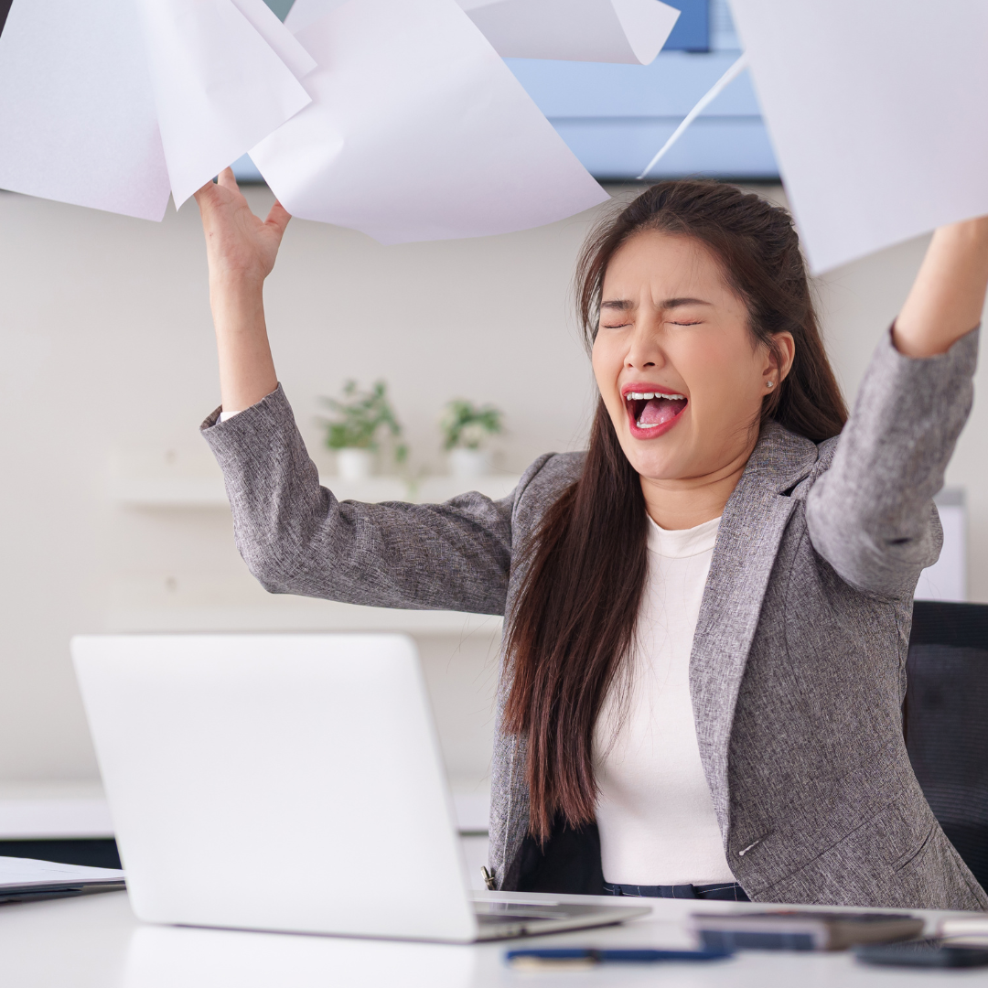 Woman sat at a desk in front of a laptop. She looks in distress and is throwing paper up in the air in frustration