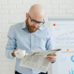 A bald man with a beard, wearing glasses and a light blue shirt, holds a cup and reads a newspaper. Behind him is a flip chart displaying financial graphs and charts against a white brick wall.