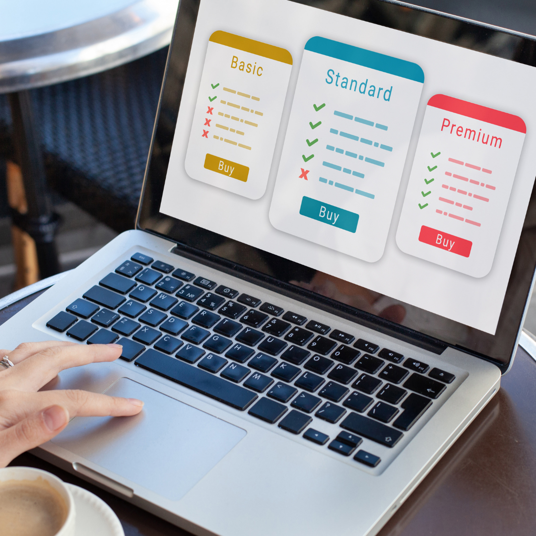 A person using a laptop displaying three pricing plans—Basic, Standard, and Premium—each with lists of features and buy buttons, on a table with a coffee cup nearby.