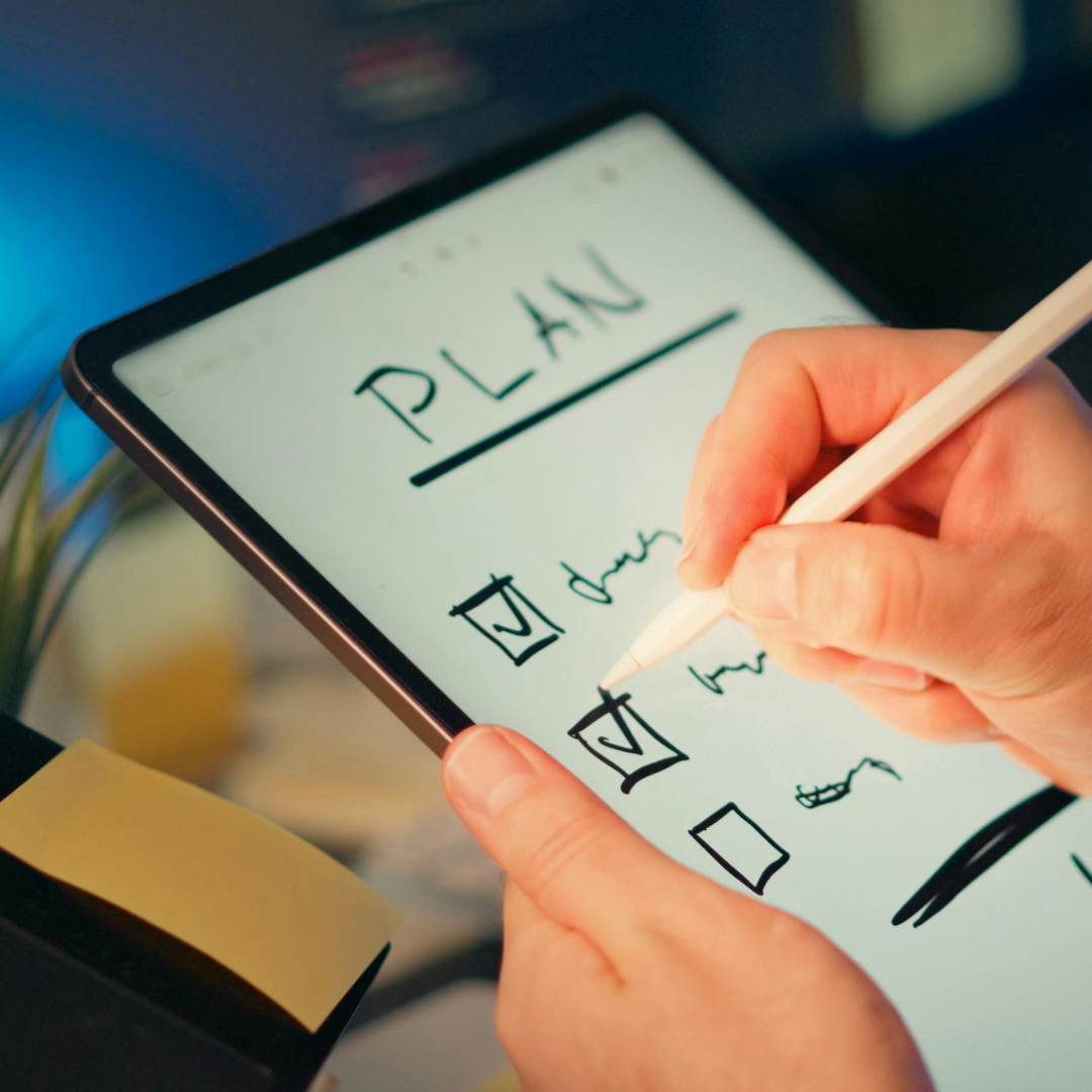 A person writes on a tablet with a stylus, marking checkboxes on a digital checklist under the heading PLAN. Sticky notes and a blurred computer screen are visible in the background.