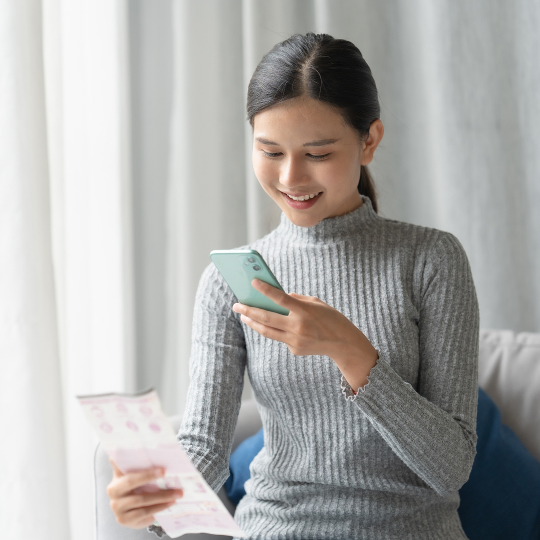 A young woman in a gray ribbed sweater smiles while looking at her smartphone and holding a piece of paper, sitting indoors on a sofa with light curtains in the background.
