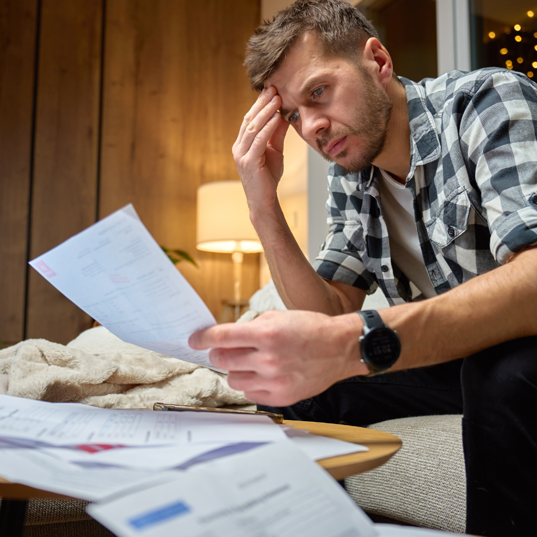 A man sits on a couch looking stressed while holding a piece of paper, surrounded by scattered documents on a table. He rests his forehead on his hand, appearing worried or overwhelmed.