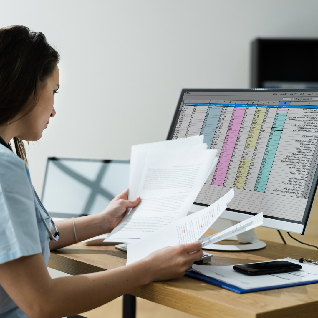 A woman sits at a desk, holding documents and reviewing papers while looking at a computer monitor displaying a colourful spreadsheet. A phone and other paperwork are on the desk.